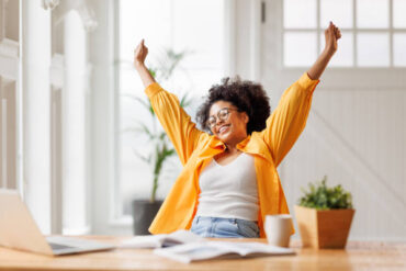 Joyful african american business woman freelancer   smiling and rejoices in victory while sitting at desk   and working at laptop screen after finishing project  in home office